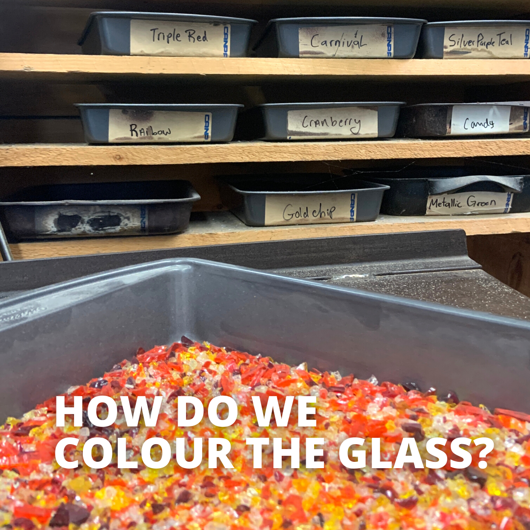 A tray of orange, red, and yellow glass frit sits on a marver in front of other trays of coloured frit in the Gray Art Glass glassblowing studio. Text reads "How do we colour the glass?"