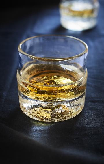 A clear crackle dip whiskey glass, filled with gold coloured whiskey, sits on a black tabletop.
